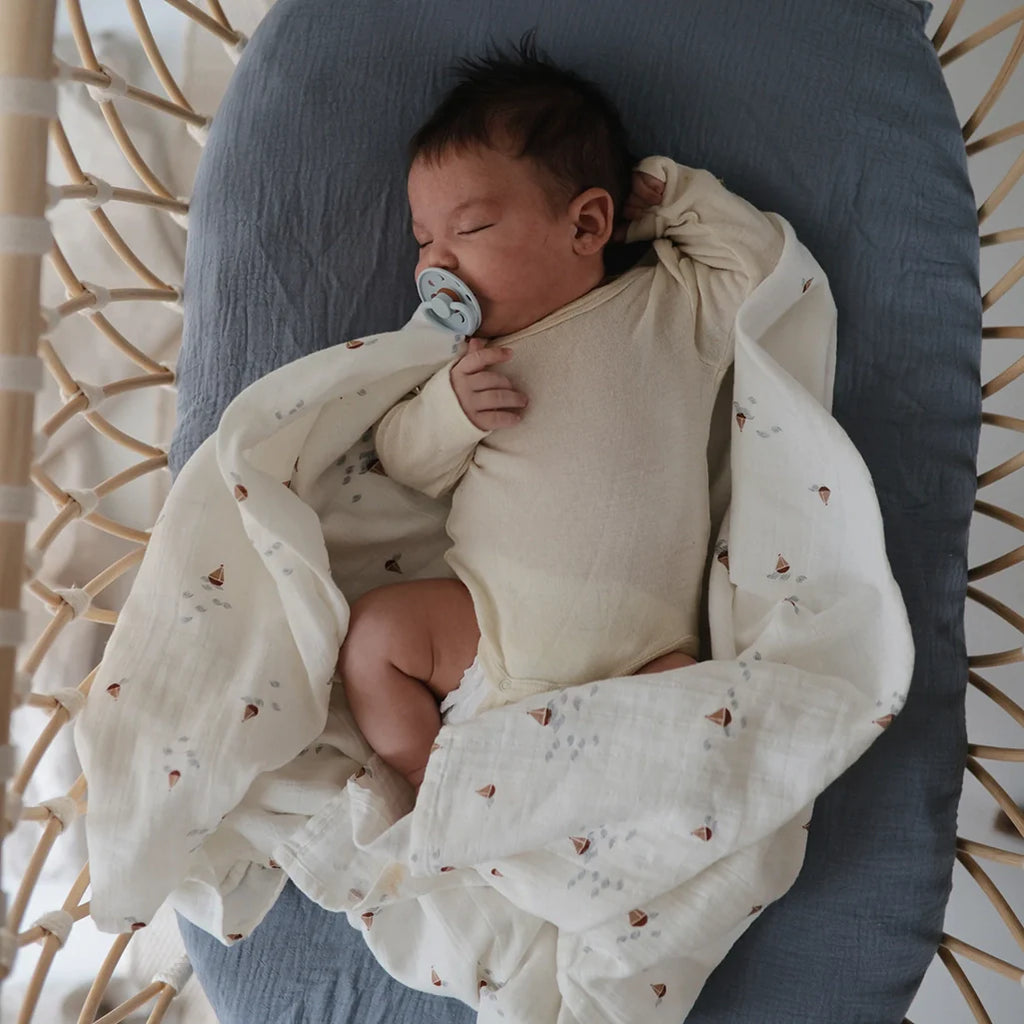 Mushie Boats Organic Cotton Nursery Swaddling Blanket. Modeled with infant in a bassinet.