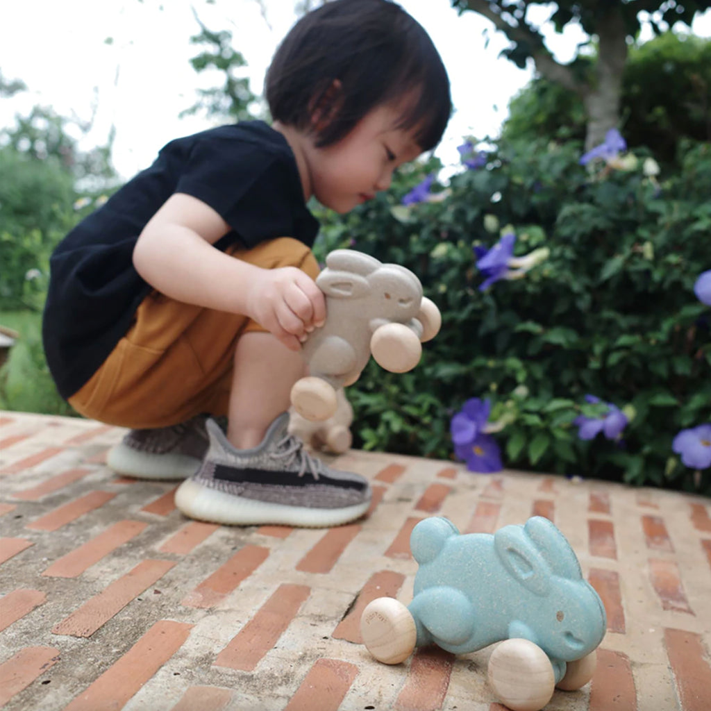 Plan Toys White Push Along Bunny Children's Active Push & Pull Toy - A small child is squatting to play while holding a white bunny in their hands. A blue bunny rests by their feet patiently 
