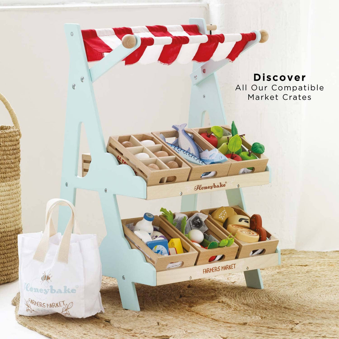 Le Toy Van's Market Stall pictured beside extra wooden crates and two cotton bags resting on a wood floor