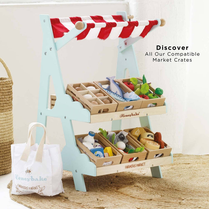 Le Toy Van's Market Stall pictured beside extra wooden crates and two cotton bags resting on a wood floor