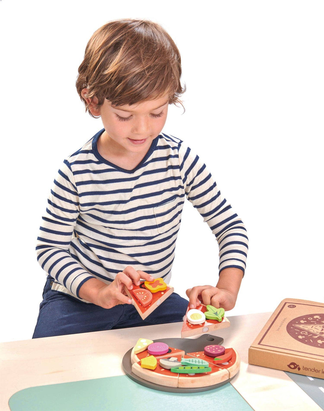 A boy in a striped shirt arranges toppings on a Tender Leaf Toys Pizza Party