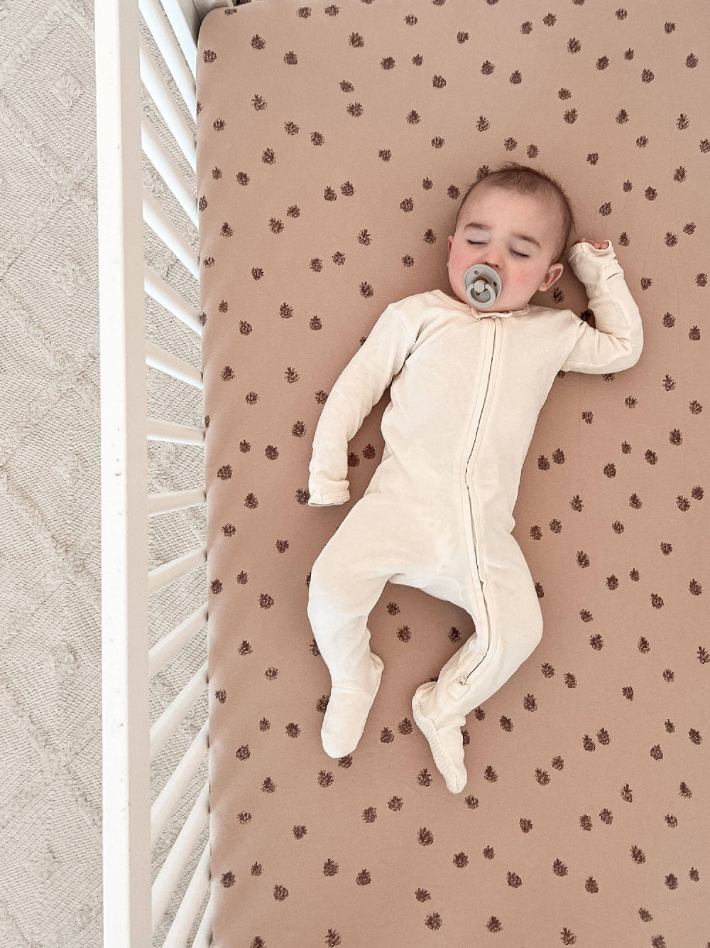 baby in white zippered footie sleeping on top organic cotton pinecone loved baby sheets sucking on green pacifier 