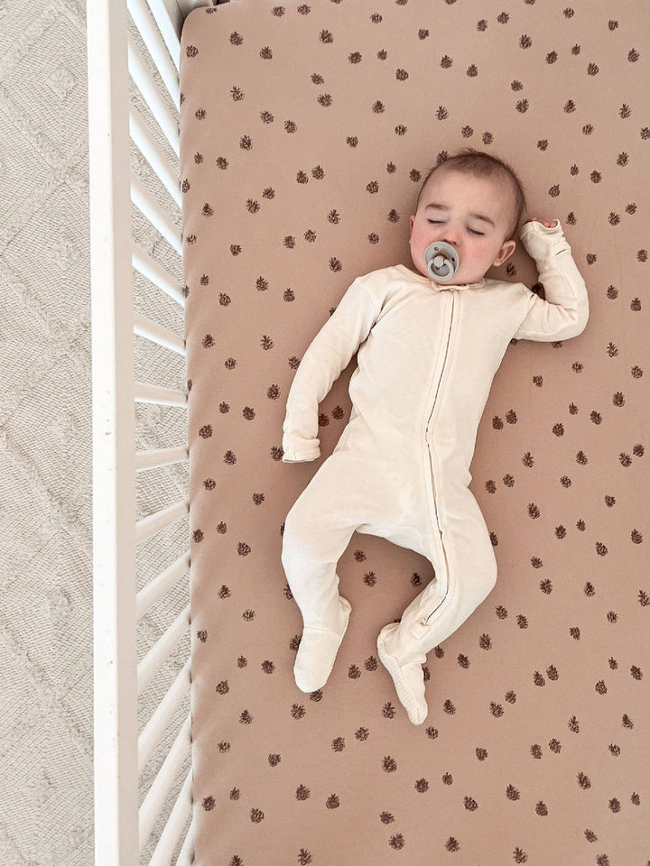 baby in white zippered footie sleeping on top organic cotton pinecone loved baby sheets sucking on green pacifier 