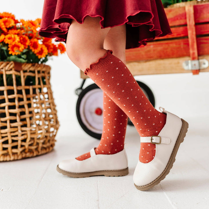 A toddler on a tricycle wears Little Stocking Co. Dottie Knee Socks in Rust with mary jane shoes