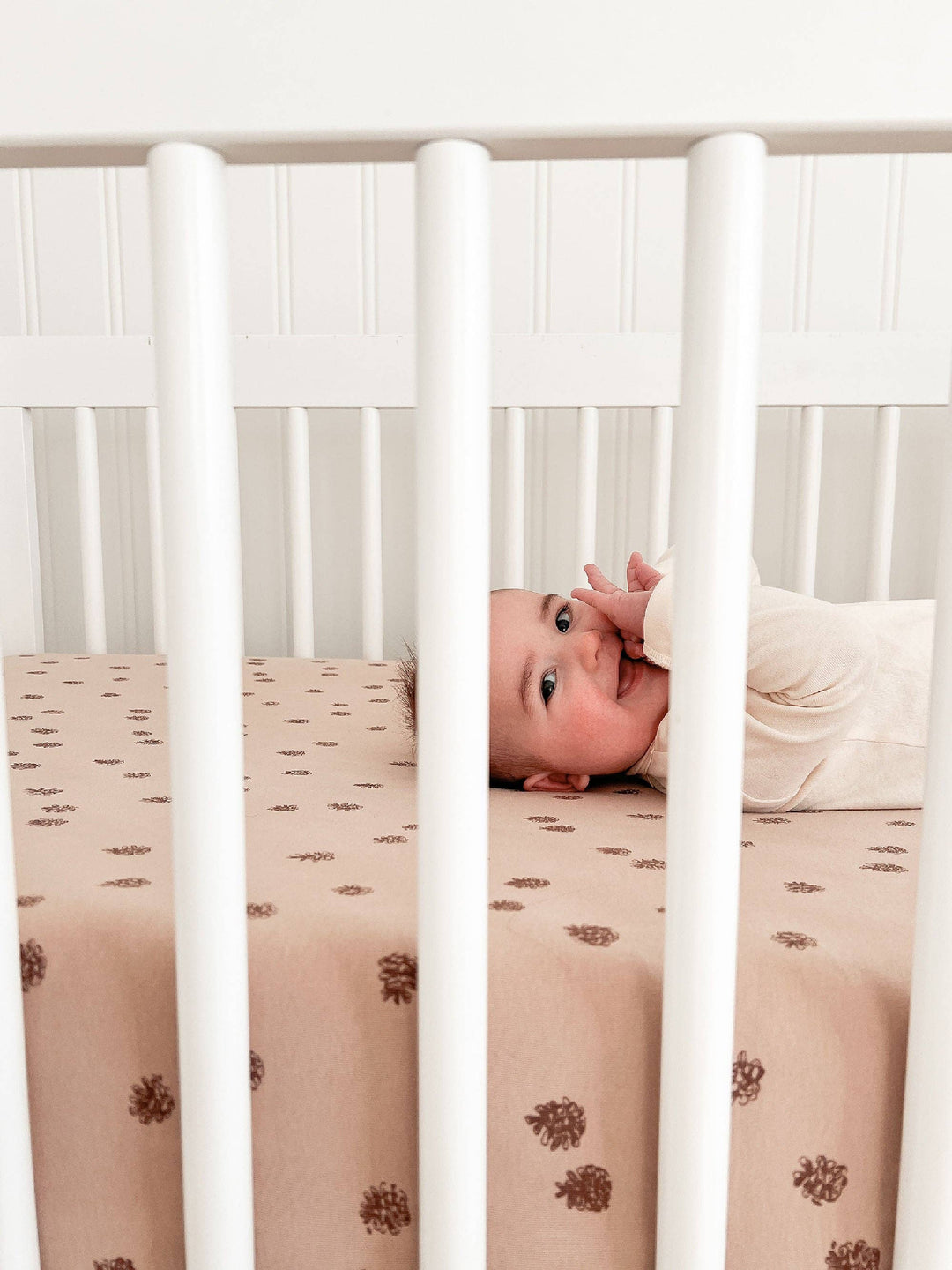 infant smiling through the bars of a minimalist white crib with loved baby oat brown crib sheets as the perfect nursery decor 