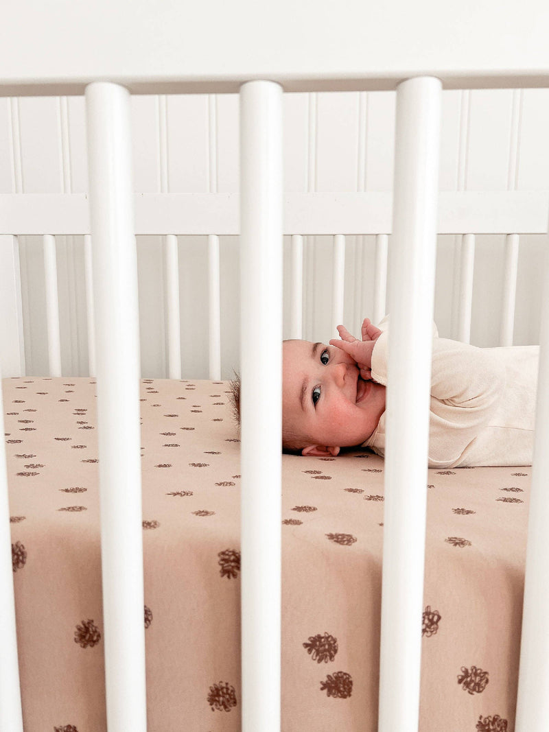 infant smiling through the bars of a minimalist white crib with loved baby oat brown crib sheets as the perfect nursery decor 