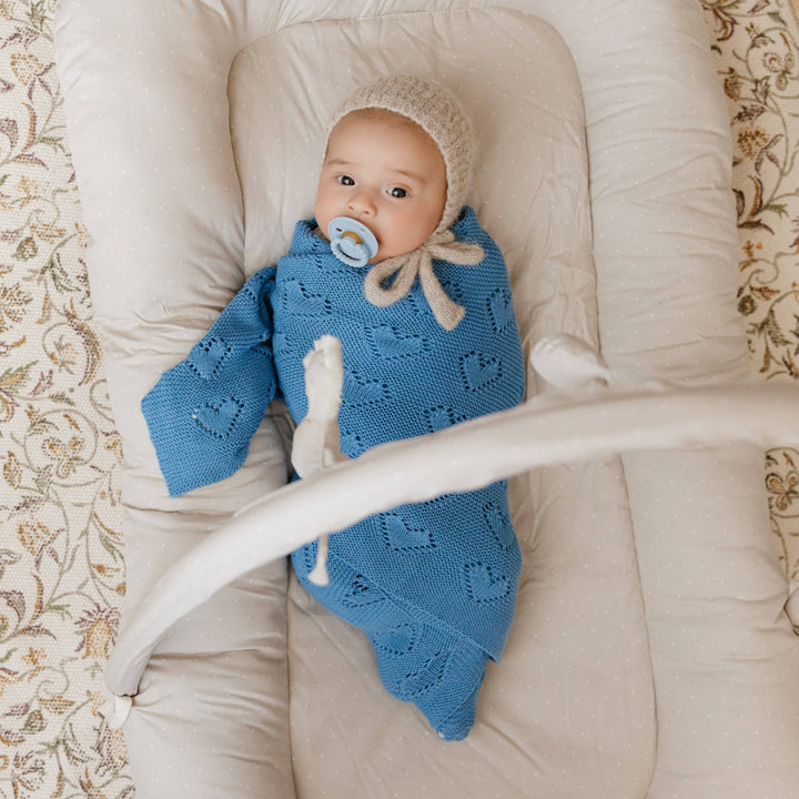 Baby wrapped in a blue knitted Bleu La La Cotton Receiving Swaddle Baby Blanket with a Heart Pattern in Denim Blue with a white bow, lying on a beige surface.