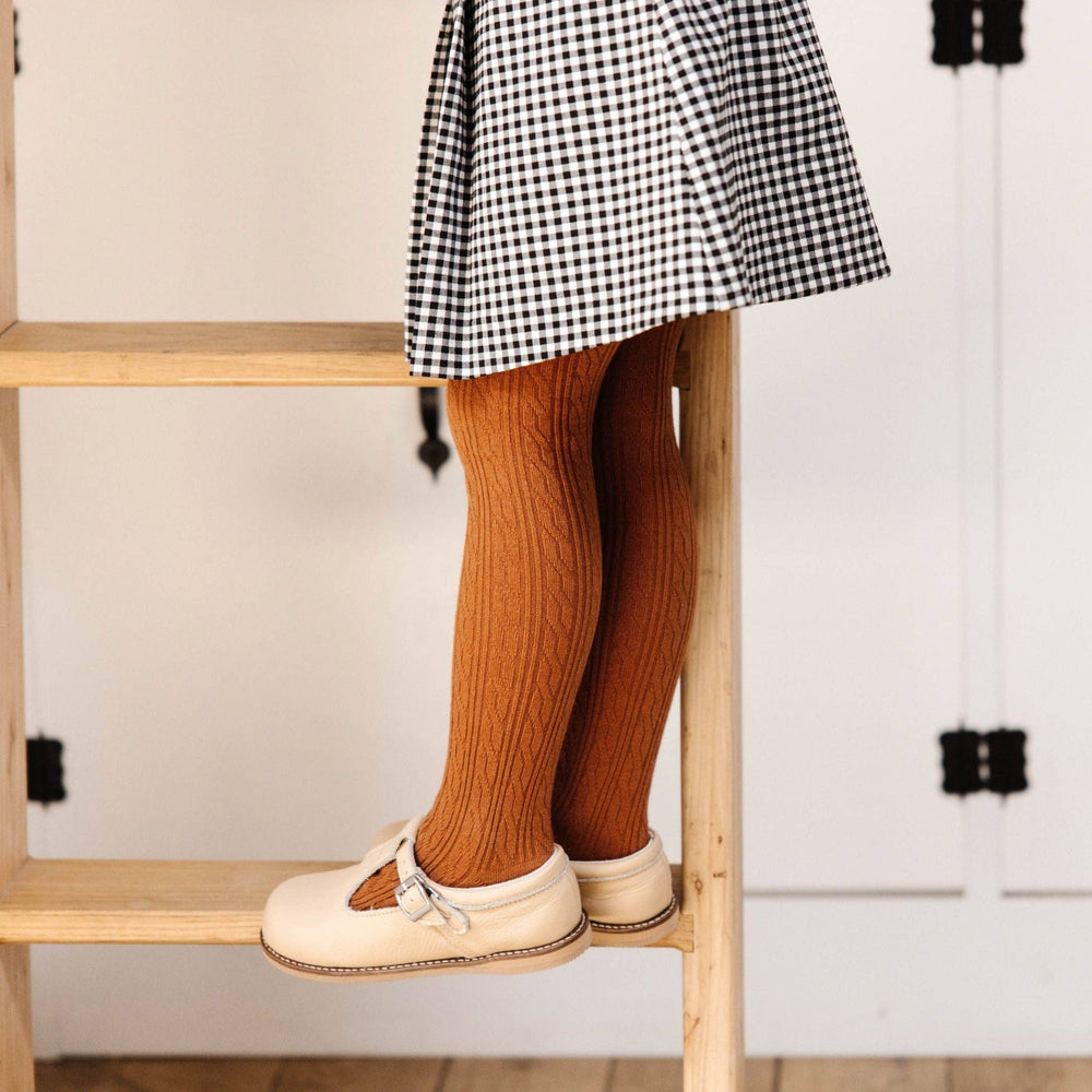 Toddler standing in Little Stocking Co. Sugar Almond Cable Knit Tights with a gingham skirt and mary janes