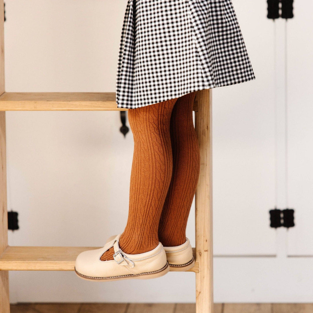 Toddler standing in Little Stocking Co. Sugar Almond Cable Knit Tights with a gingham skirt and mary janes