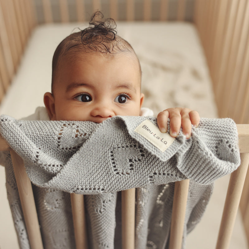 Baby standing in crib wrapped in Bleu La La 100% Cotton Luxury Knit Heart Swaddle Baby Blanket in grey for everyday soothing.