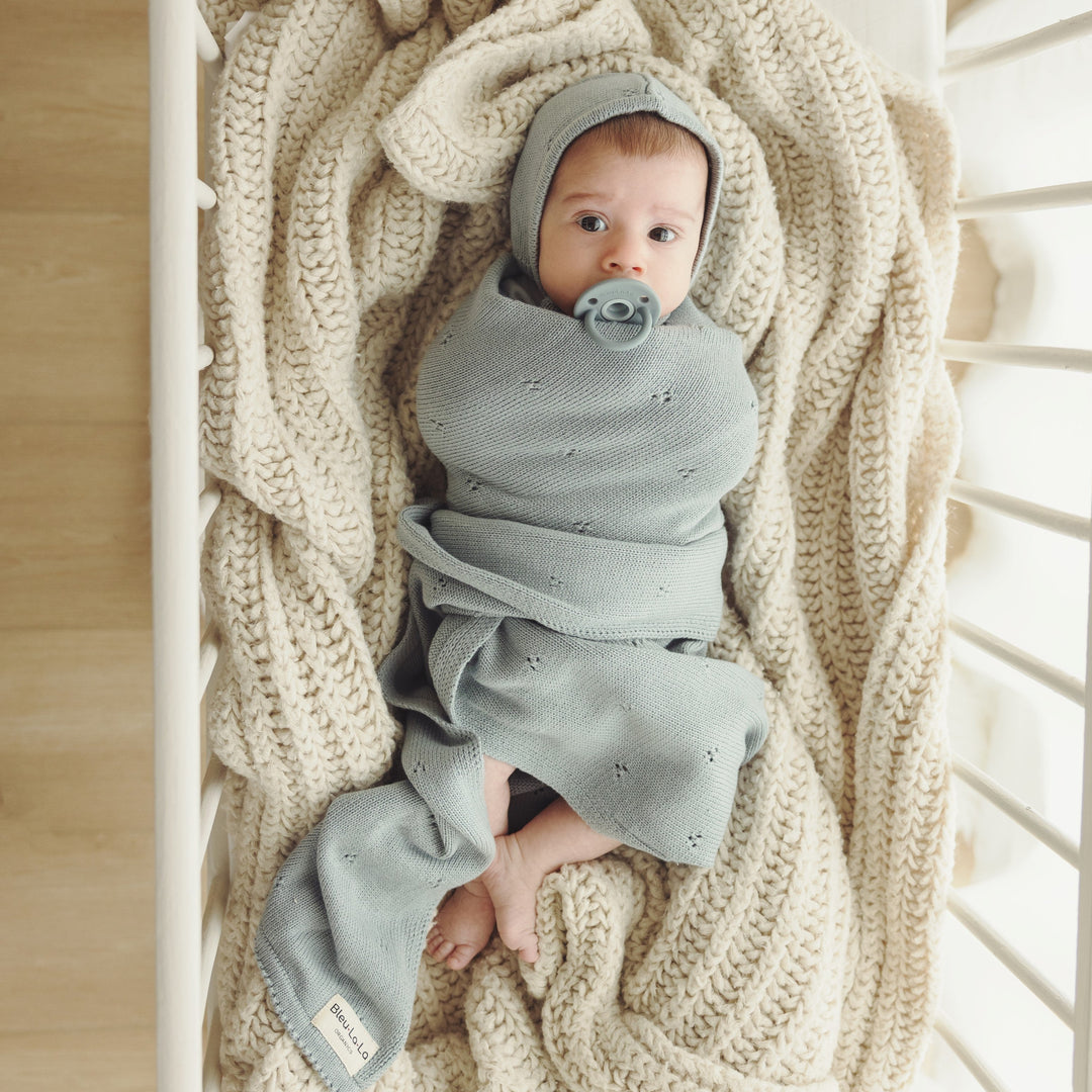 Baby swaddled in Bleu La La Organic Pointelle Swaddle Blanket in powder blue inside a crib, highlighting soft organic cotton knit.