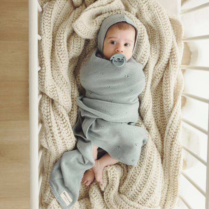 Baby swaddled in Bleu La La Organic Pointelle Swaddle Blanket in powder blue inside a crib, highlighting soft organic cotton knit.