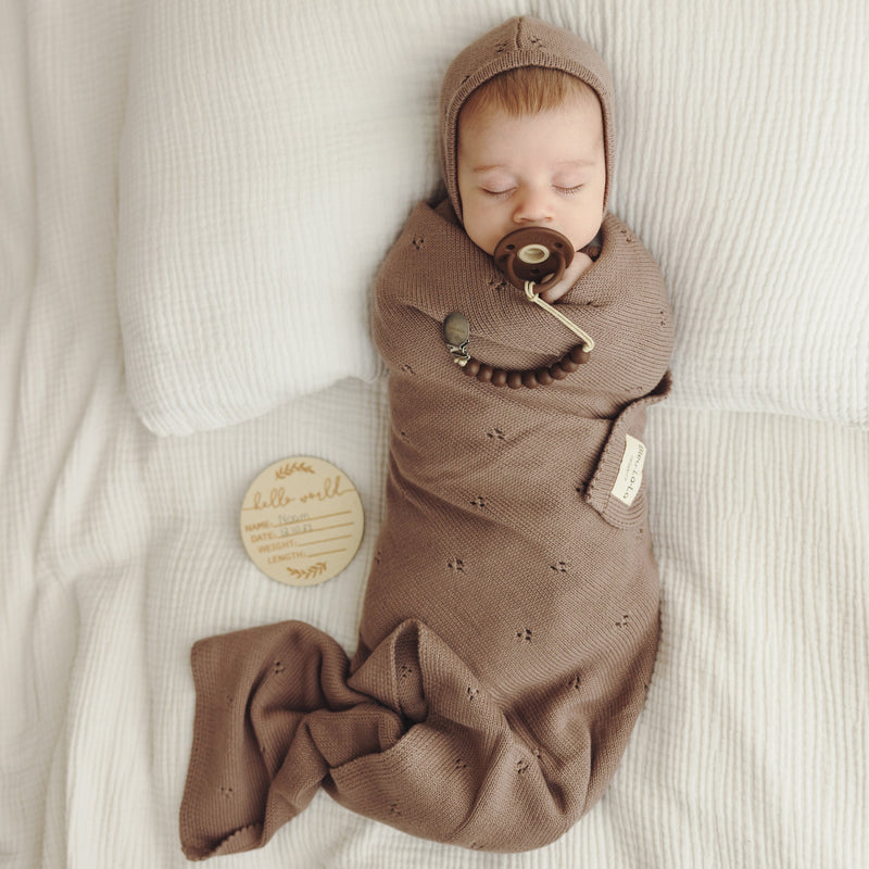 Newborn sleeping swaddled in Bleu La La Organic Pointelle Swaddle Blanket in mocha with coordinating organic cotton hat.