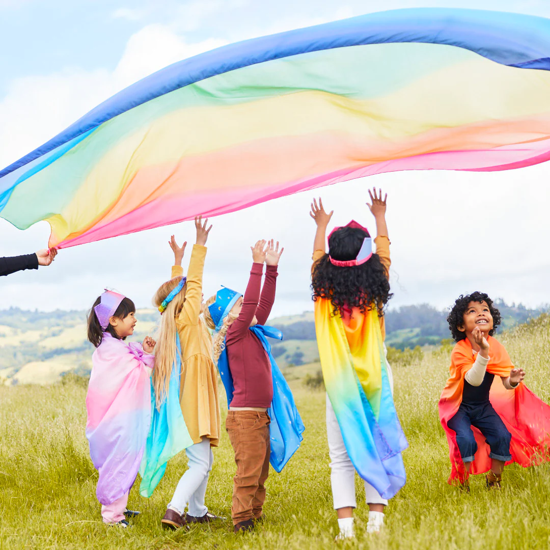 Kids run outdoors holding a rainbow silk overhead, playing parachute games with Sarah's Silks