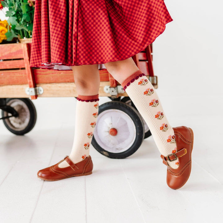 Toddler in a red dress wearing Little Stocking Co. Posie Knee Socks in Rust with mary jane shoes