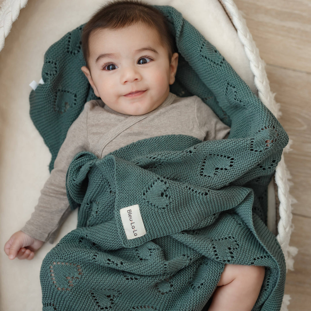 Baby wrapped in a green knitted Bleu La La Cotton Receiving Swaddle Baby Blanket with a Heart Pattern in sage, sitting in a white basket.