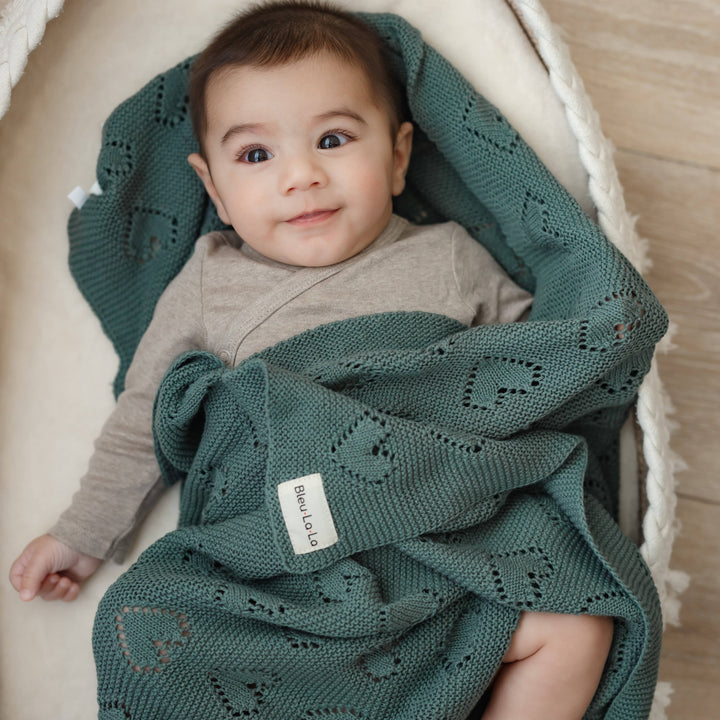 Baby wrapped in a green knitted Bleu La La Cotton Receiving Swaddle Baby Blanket with a Heart Pattern in sage, sitting in a white basket.