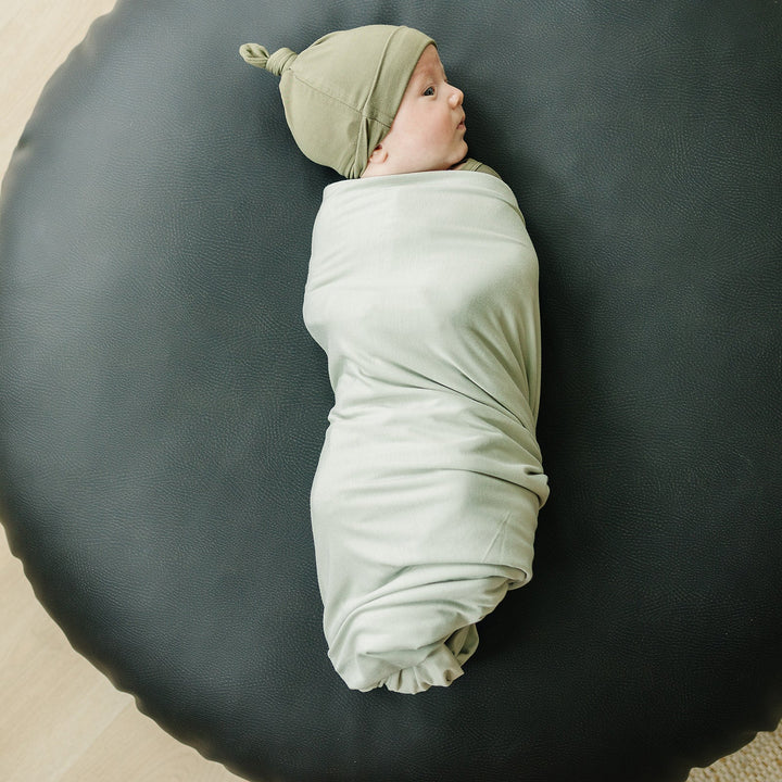 Sage Bamboo Stretch Swaddle draped over a crib rail, a soft stretchy receiving blanket for baby boys and girls.