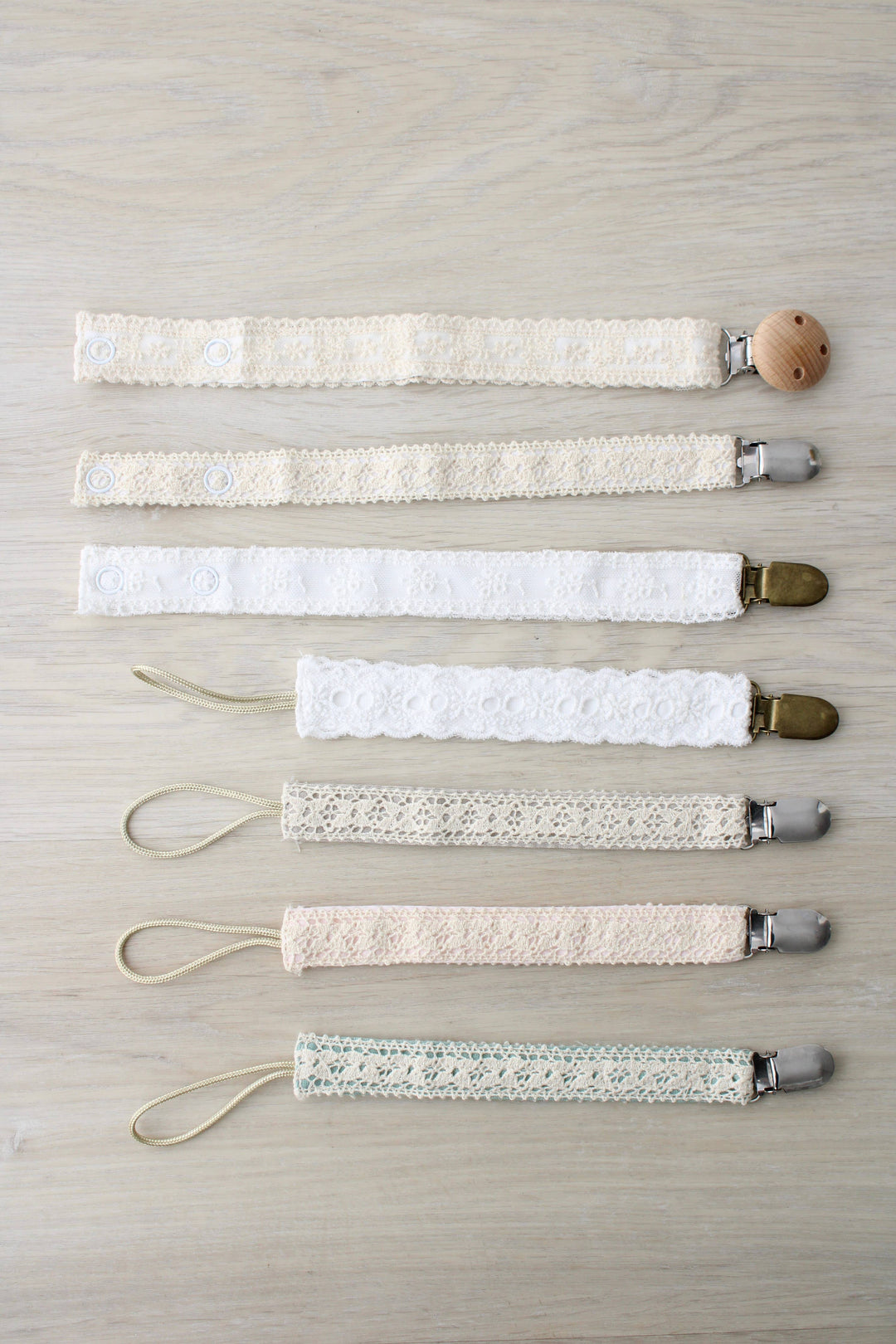 Seven cream and ivory paci clips in a row, including the Little Threads Ivory Floral Lace Pacifier Clip