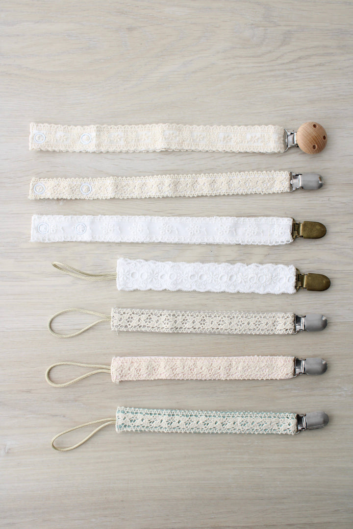 Seven cream and ivory paci clips in a row, including the Little Threads Ivory Floral Lace Pacifier Clip