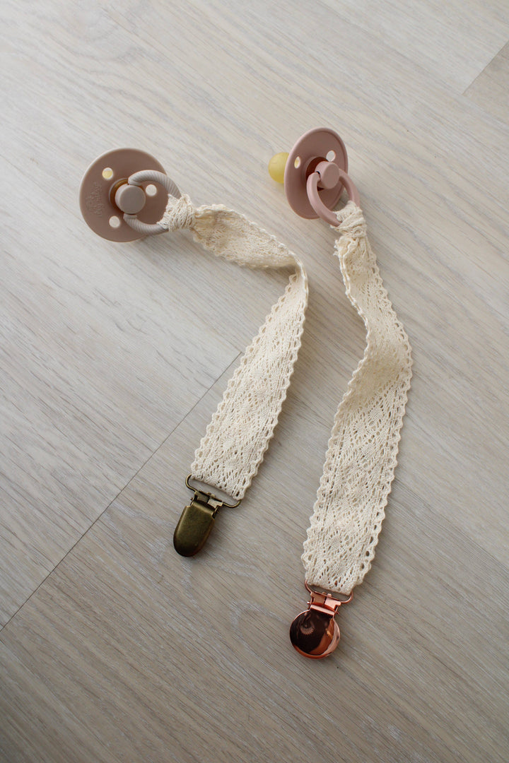 Cream crochet pacifier holders with pacifiers attached on a wood floor, Little Threads Diamond Crochet Collection clips