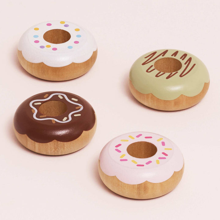 Four wooden donuts in vanilla, pistachio, chocolate, and strawberry glazes sit atop a blush pink surface