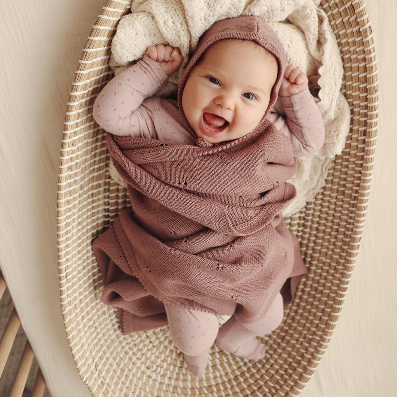 Baby wrapped in a 100% Organic Cotton Pointelle Swaddle Receiving Baby Blanket in Sugar Plum Purple with a bonnet, smiling in a woven basket.