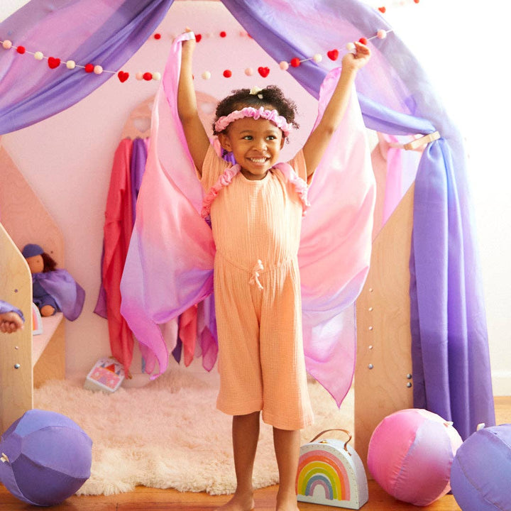 Arms raised high, a joyful child celebrates in the Sarah's Silks Blossom Garland at playtime