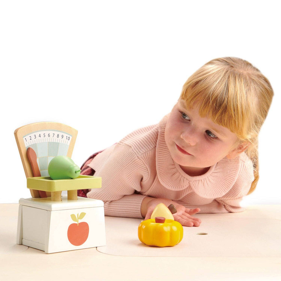 A little girl leans close to examine the Tender Leaf Toys Market Scales with fruit nearby
