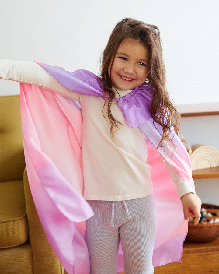 At home, a child models the Sarah's Silks Blossom Cape with arms stretched outward