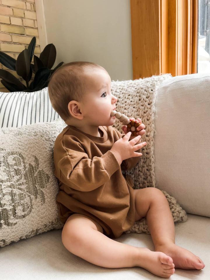 baby in rusty red boho and babe bubble romper sitting on linen couch and chewing on natural wood teether toy 