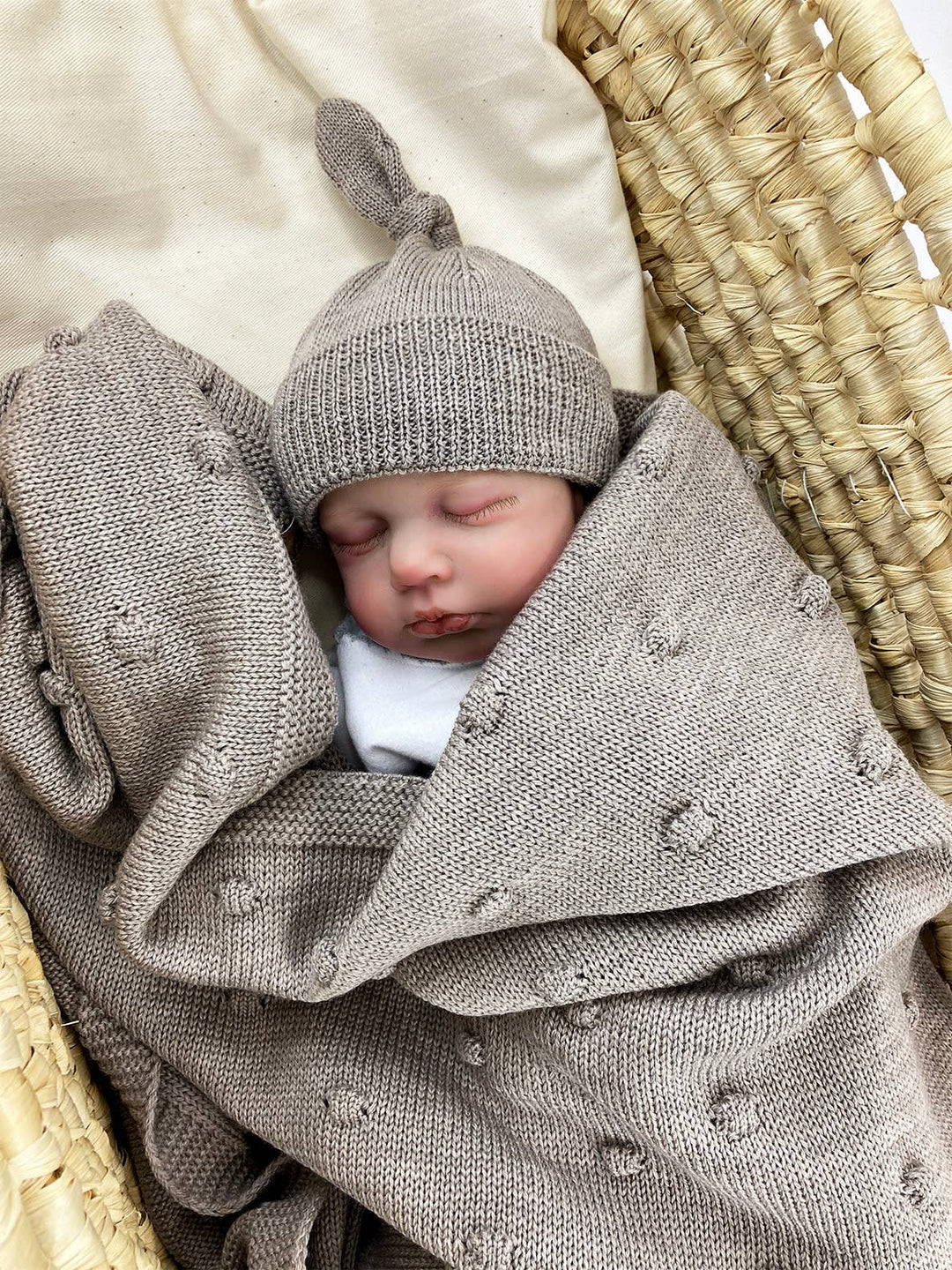 Sleeping baby in a wicker basket wearing the beige ALIAP Merino Wool Top Knot Baby Beanie with a matching knit blanket