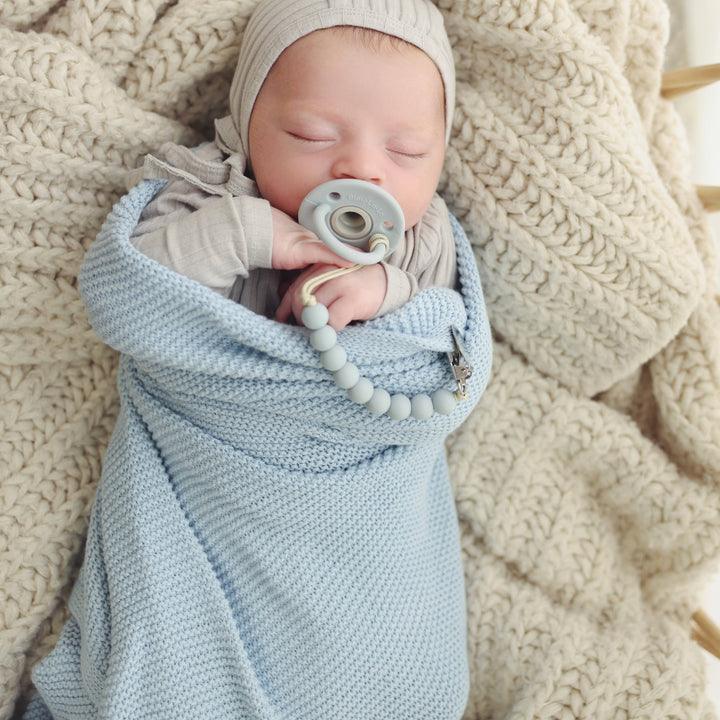 Newborn baby wrapped in a Bleu La La Heritage Knit 100% Organic Cotton Swaddle Baby Blanket with a pacifier, lying on a textured beige blanket