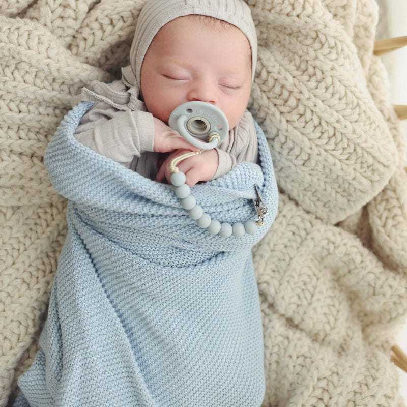 Newborn baby wrapped in a Bleu La La Heritage Knit 100% Organic Cotton Swaddle Baby Blanket with a pacifier, lying on a textured beige blanket