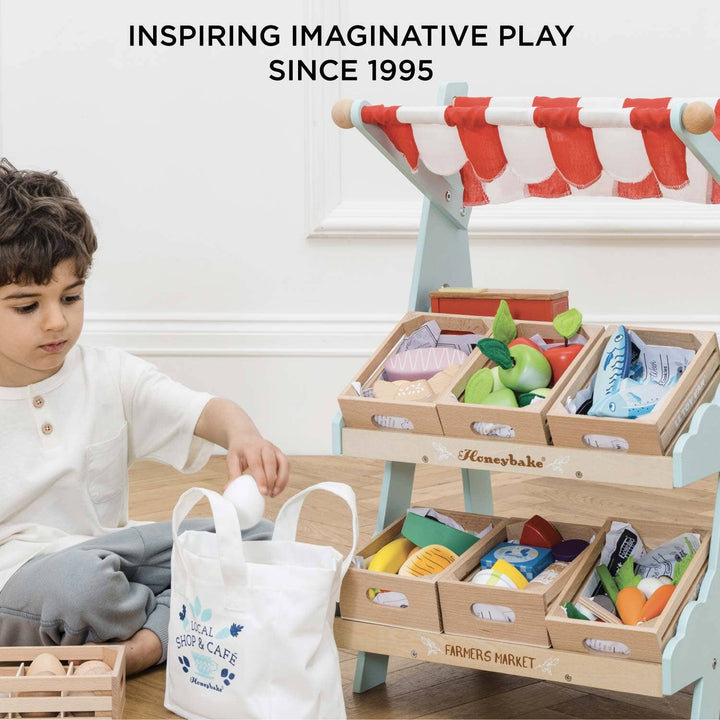 A toddler reaches into a wooden crate of pretend produce on the Le Toy Van Market Stall shelf