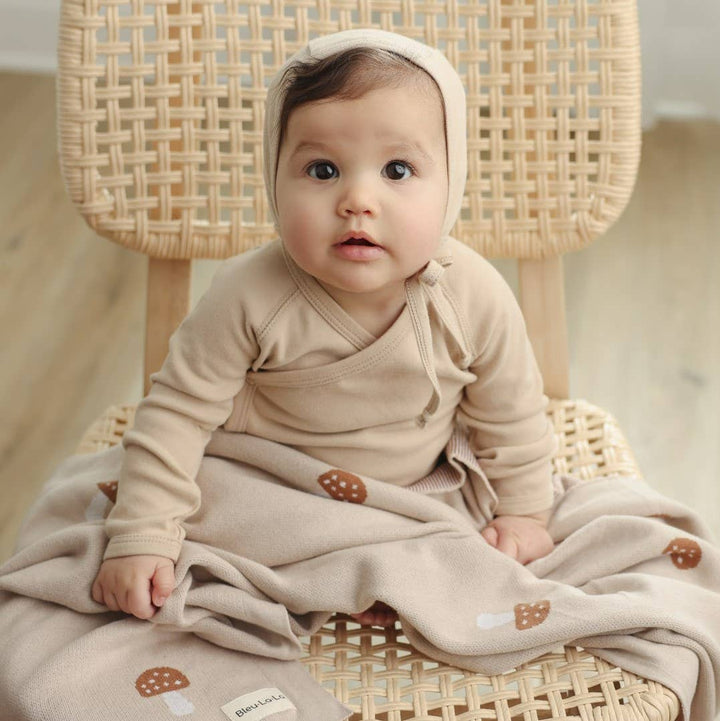 Baby in a beige outfit sitting on a wicker chair with a soft Bleu La La Cotton Swaddle Receiving Mushroom Blanket in Camel