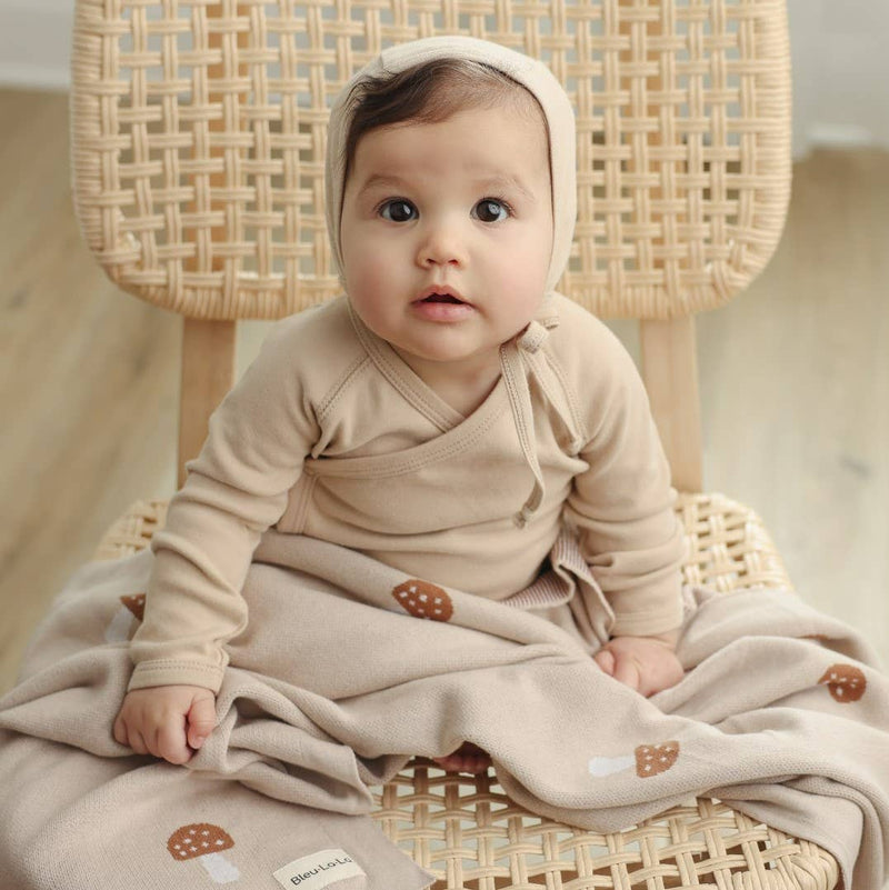 Baby in a beige outfit sitting on a wicker chair with a soft Bleu La La Cotton Swaddle Receiving Mushroom Blanket in Camel