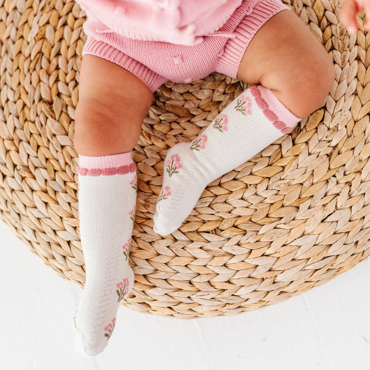 Toddler sitting on a woven rug wearing Little Stocking Co. Posie Knee Socks in Old Rose, ruffle-top baby socks