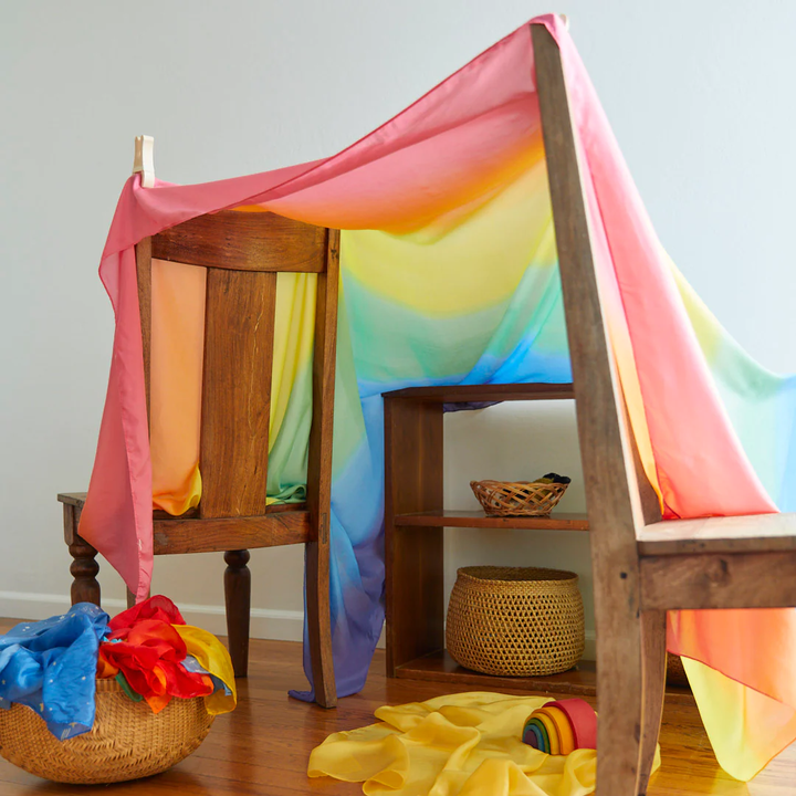 A wooden chair becomes a cozy play fort draped in the Double Rainbow Playsilk from Sarah's Silks