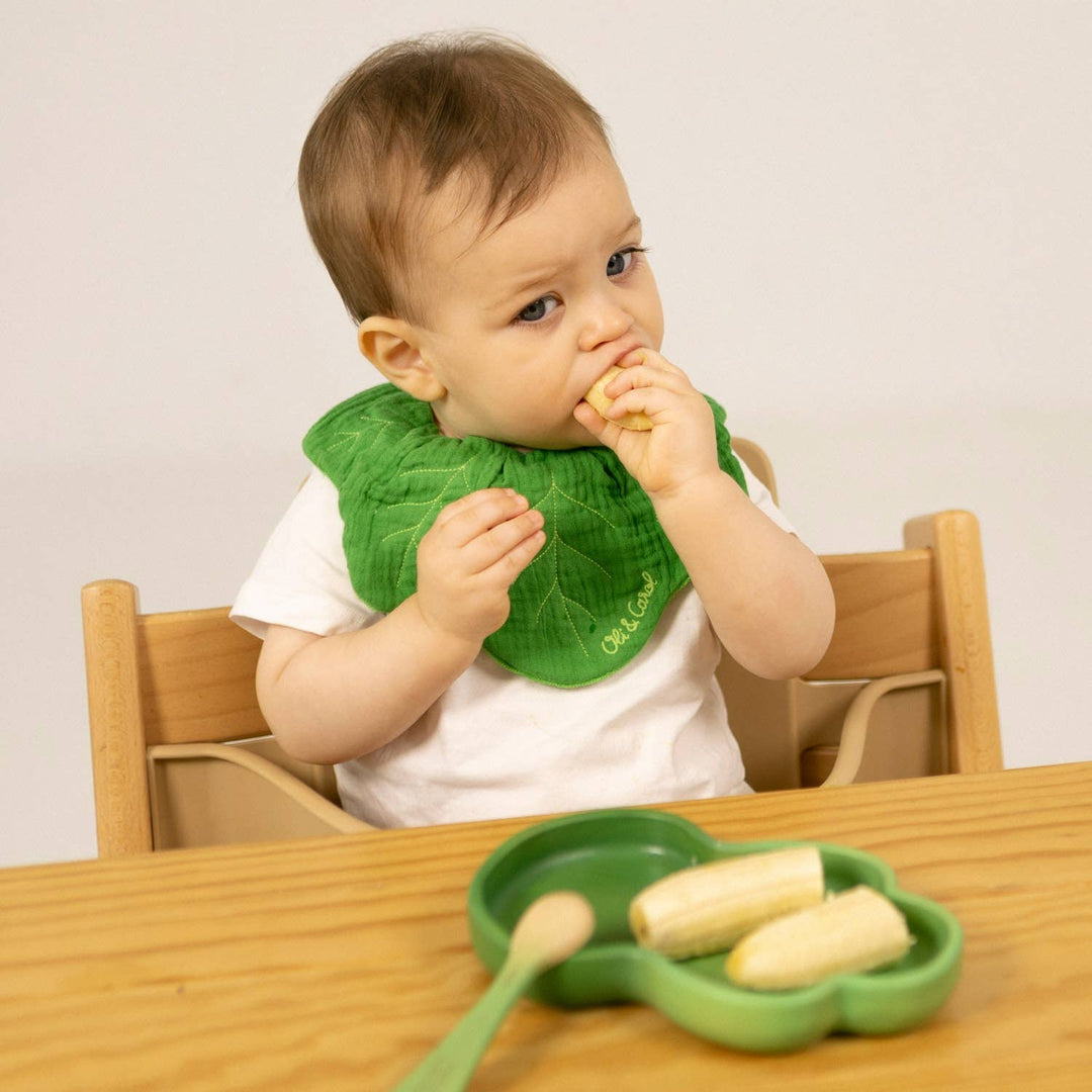 A baby in a bib feeds himself using the Oli & Carol Kendall the Kale Plate & Spoon Set