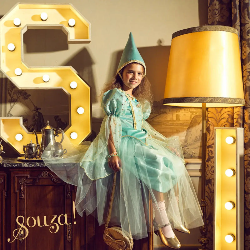 Child in a teal costume and Souza Victoria Medieval Hat sitting on a chair with decorative letters and lamp in the background