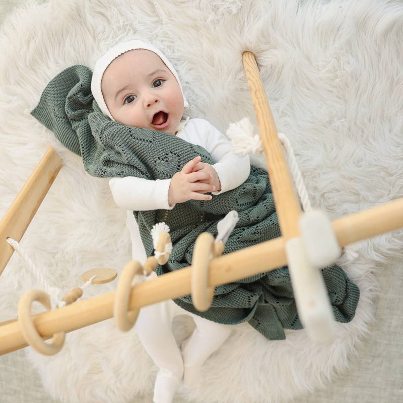 Baby in a green knitted Bleu La La Cotton Receiving Swaddle Baby Blanket with a Heart Pattern in sage and white headband sitting on a wooden toy with white rings on a fluffy white surface.