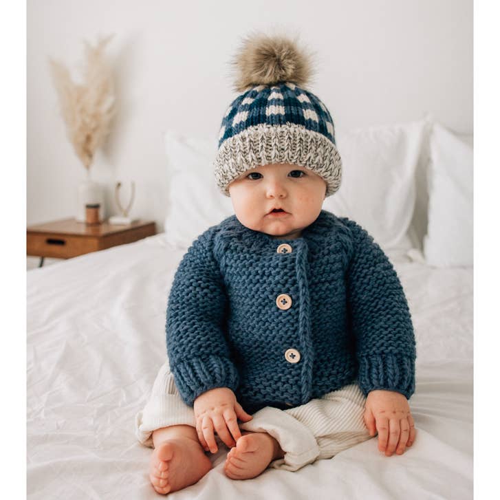Baby in Slate blue garter stitch cardigan and matching beanie by Huggalugs, sitting on bed