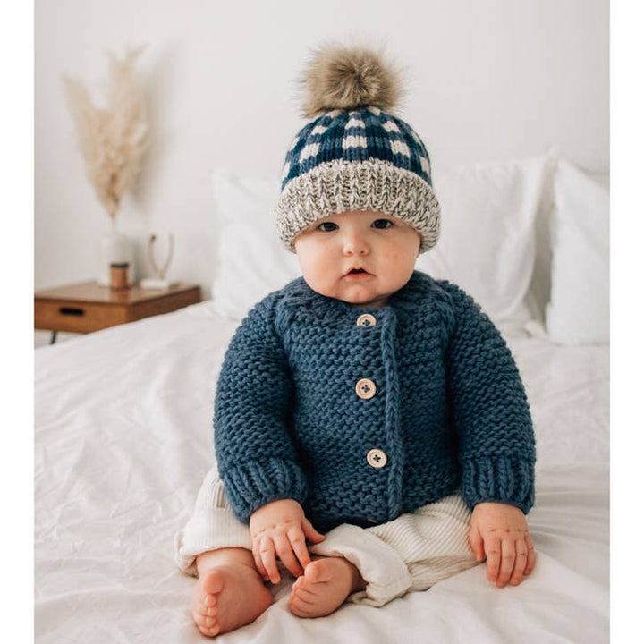 Baby in Slate blue garter stitch cardigan and matching beanie by Huggalugs, sitting on bed