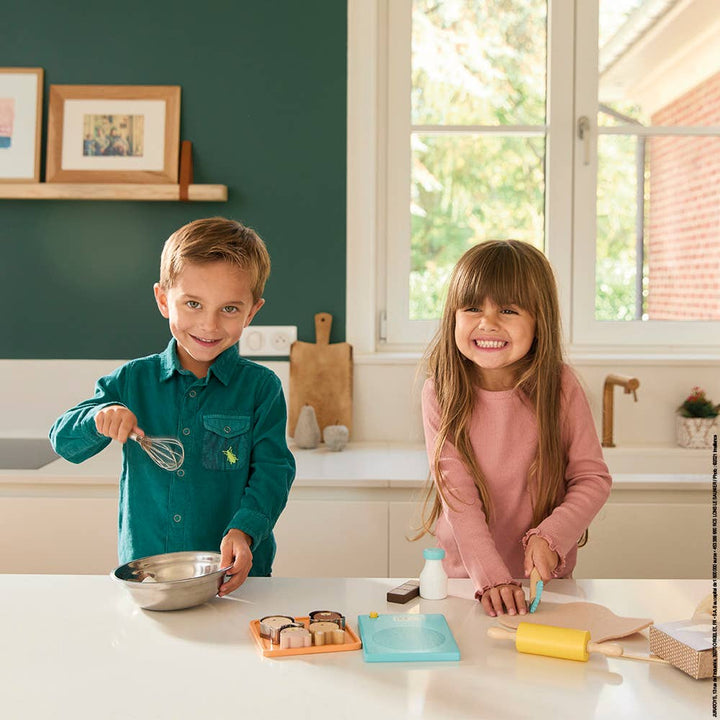 Two children in a play kitchen, one holding a whisk and the other with pretend play baking tools, smiling at the camera.
