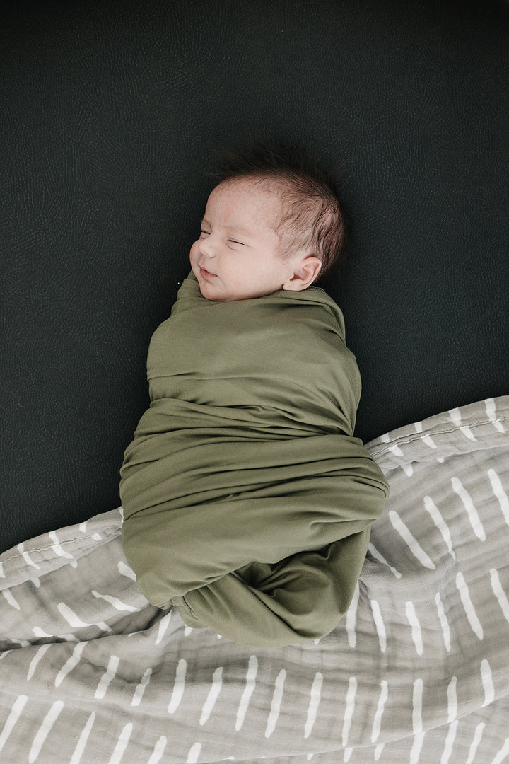 Olive Bamboo Stretch Swaddle draped over a crib rail, a breathable bamboo swaddle blanket for newborns.