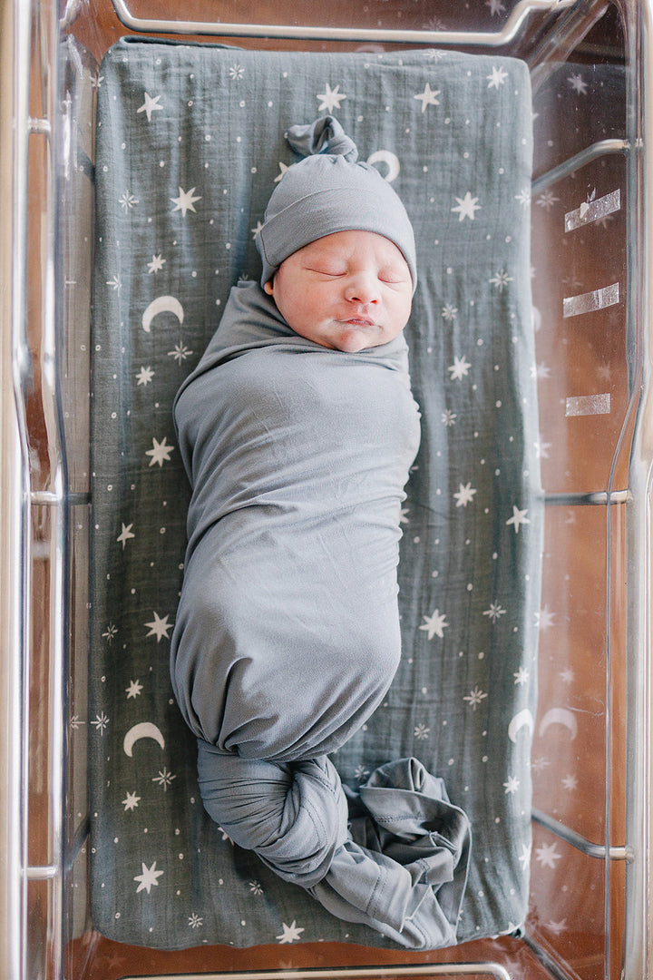 Dusty Blue Bamboo Stretch Swaddle draped over a crib rail, a breathable bamboo swaddle blanket for newborns.