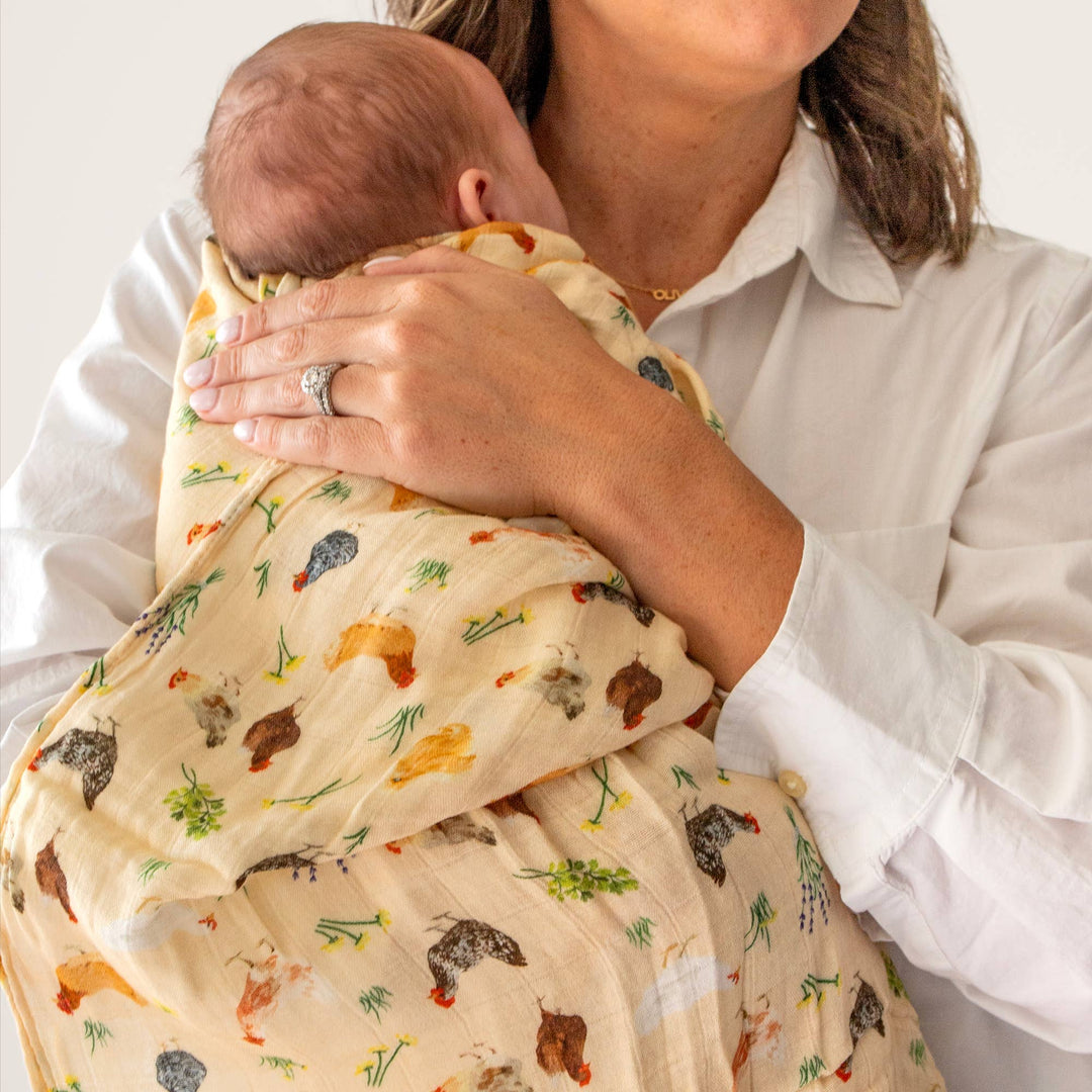 Mother swaddling newborn in 1canoe2 Chicken Meadows baby swaddle, a soft muslin blanket with chicken print