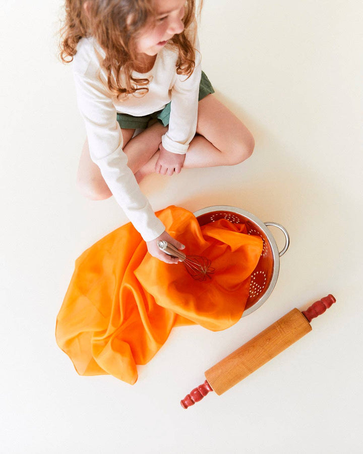 Child pretend baking with an orange Sarah's Silks Solid Color Playsilk, treasured by young children