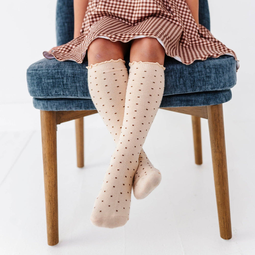 Seated on a blue chair, this toddler models Little Stocking Co. Dottie Knee Socks in Vanilla with a plaid skirt